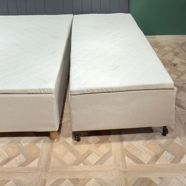 Deux modules de lit entièrement assemblés sont placés côte à côte sur un parquet clair, séparés par un étroit espace. Chaque module possède sa propre housse beige, son propre matelas matelassé et ses propres pieds.